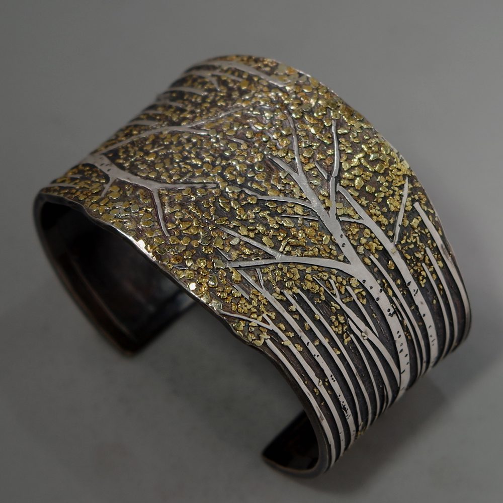 A silver cuff bracelet with trees and gold leaf.